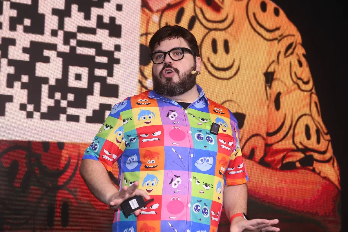 Bruno UAU com camisa colorida de smileys diante de um painel de carinhas felizes