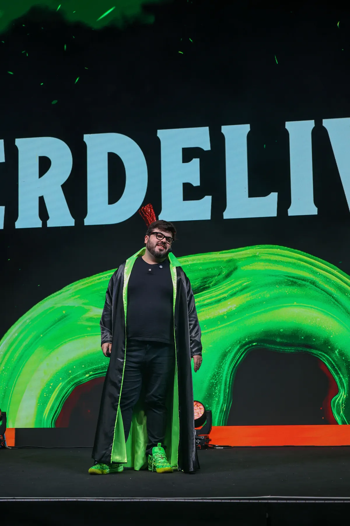 Bruno UAU de capa de vilão no palco, diante de uma maçã envenenada verde gigante no telão