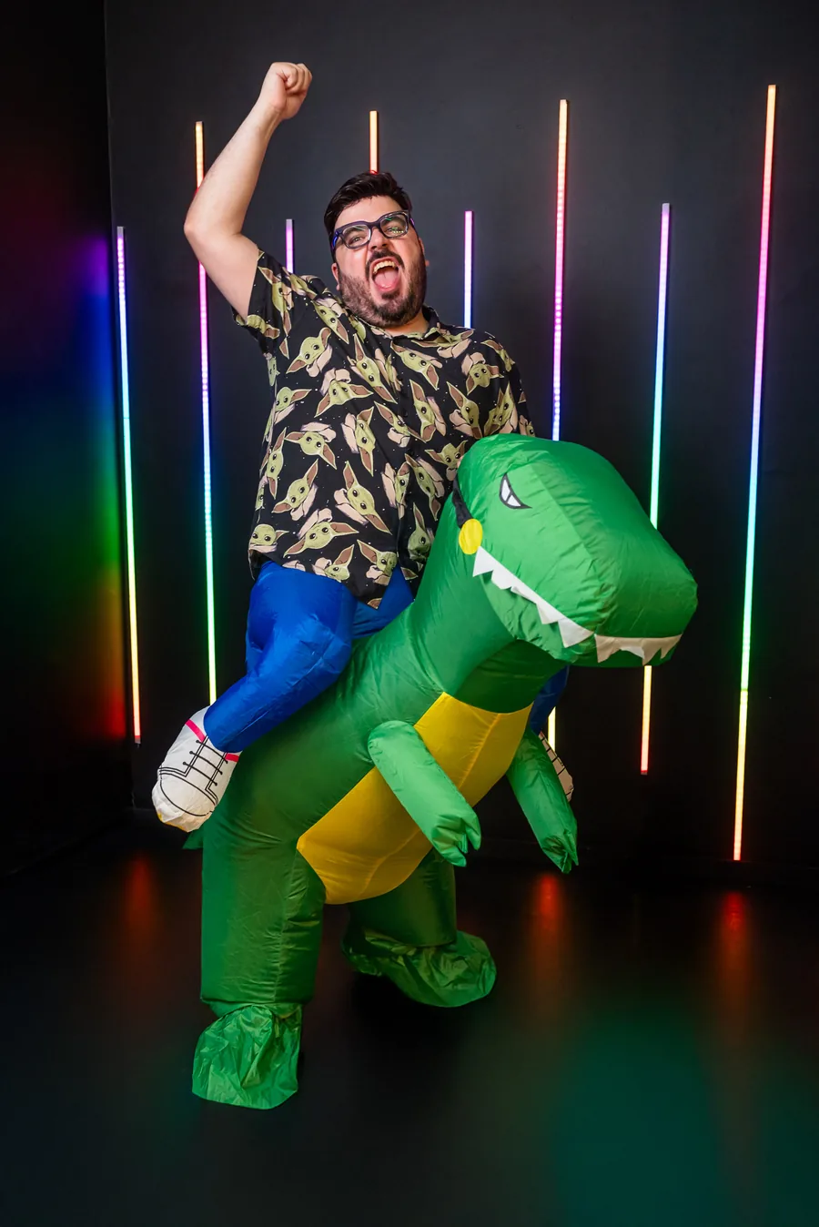 Bruno UAU montado no dinossauro inflável, de punho para cima e língua de fora, diante de luzes neon