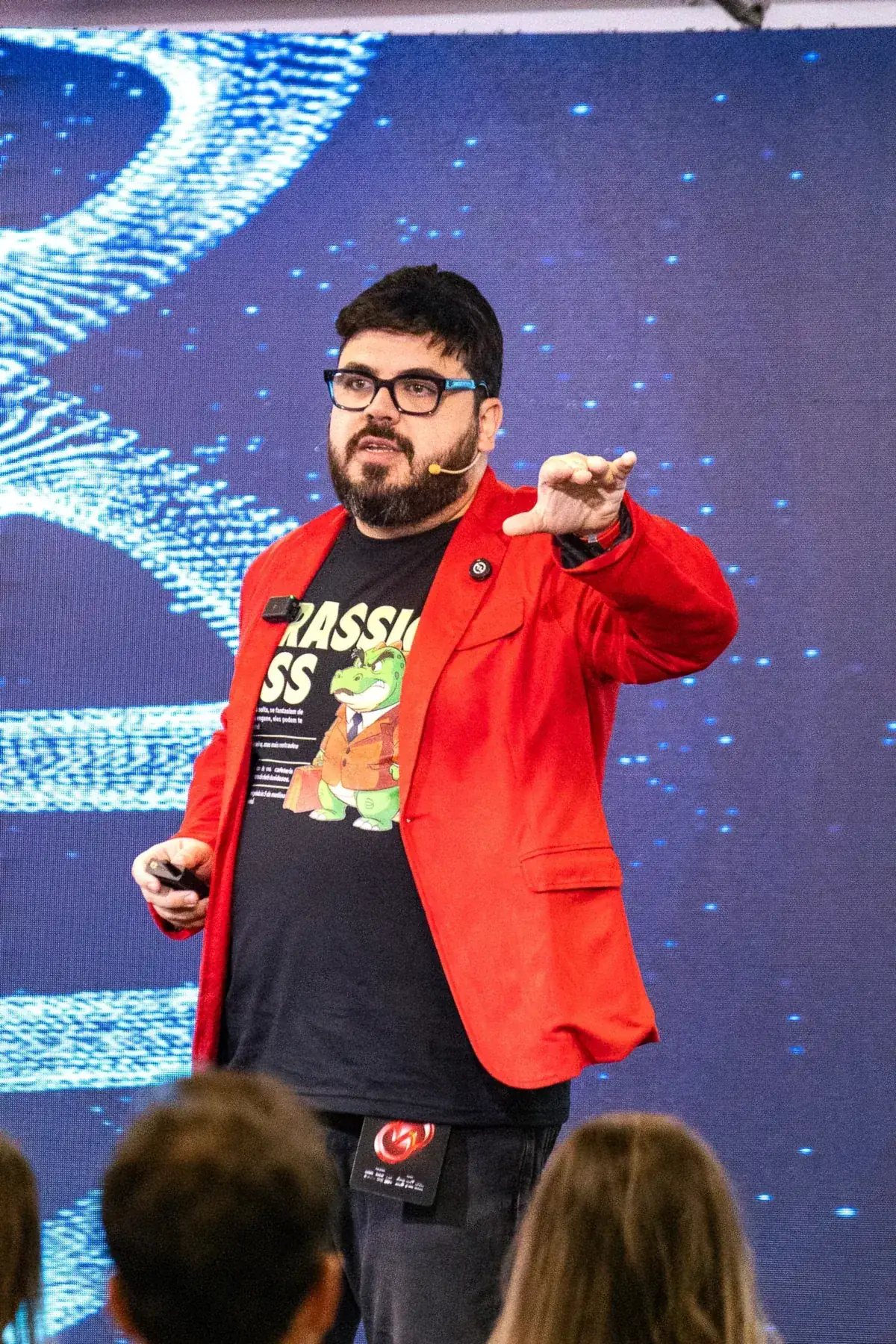 Bruno UAU de blazer vermelho e camiseta do Jurassic Boss, apontando para a plateia